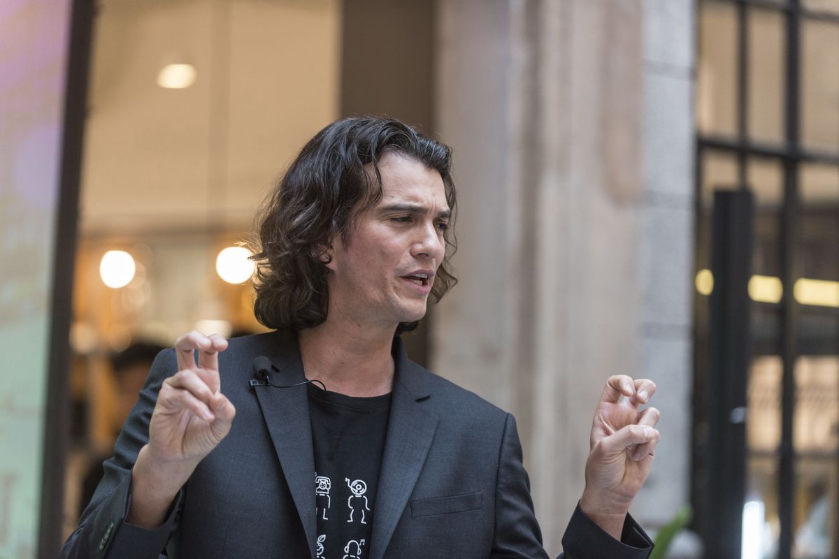 A man with shoulder-length hair wearing a t-shirt and suit jacket speaks to an off-camera crowd.