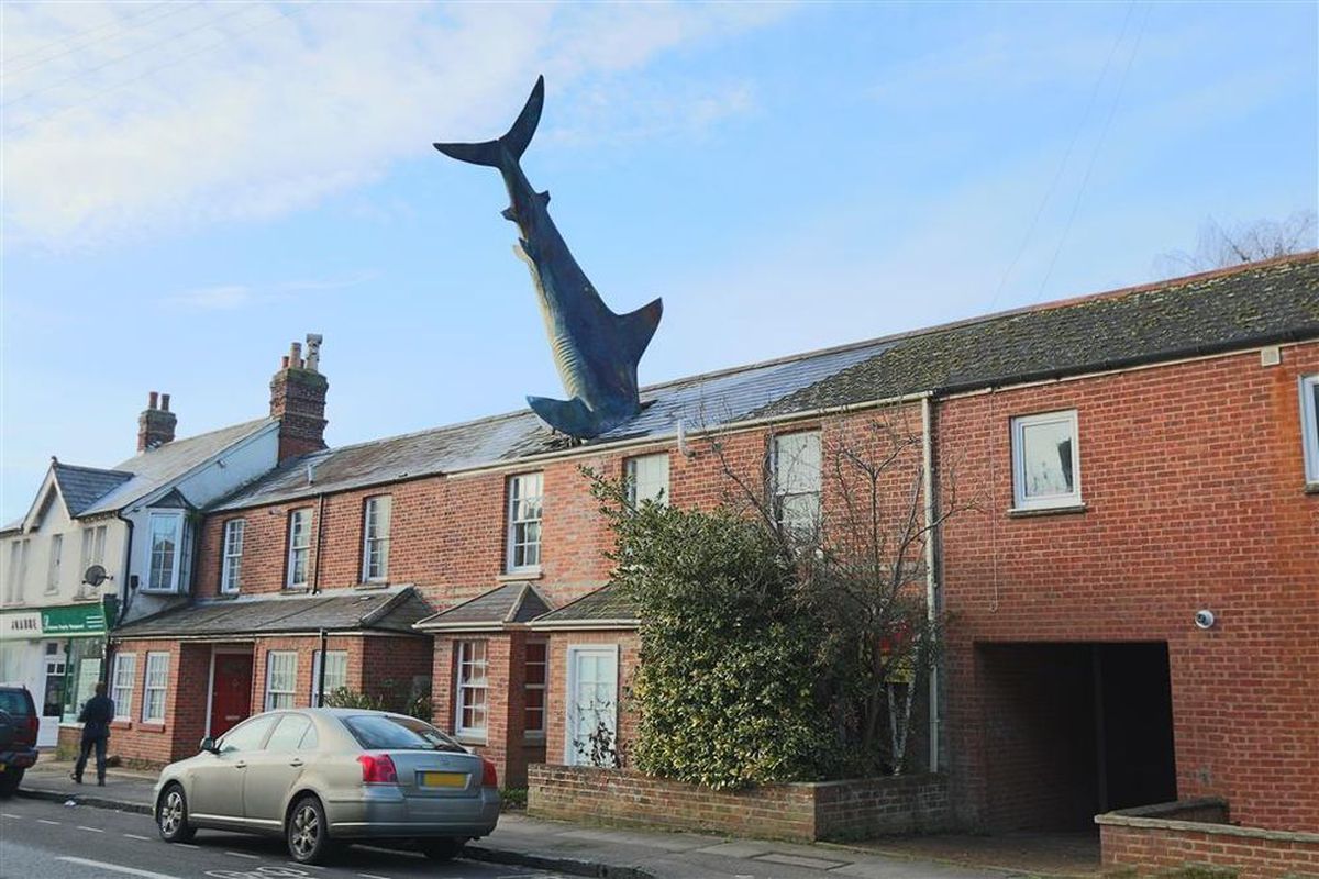 A large, fake shark sticks out of the roof of a normal-looking brick row house. 