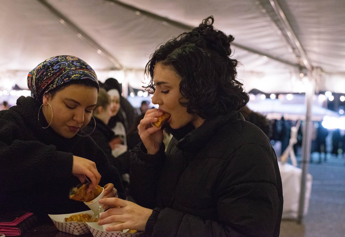 Two festival-goers, one wearing a headscarf and big hoop earrings and both wearing black jackets eating tacos at the Ramadan Suhoor Festival in Dearborn