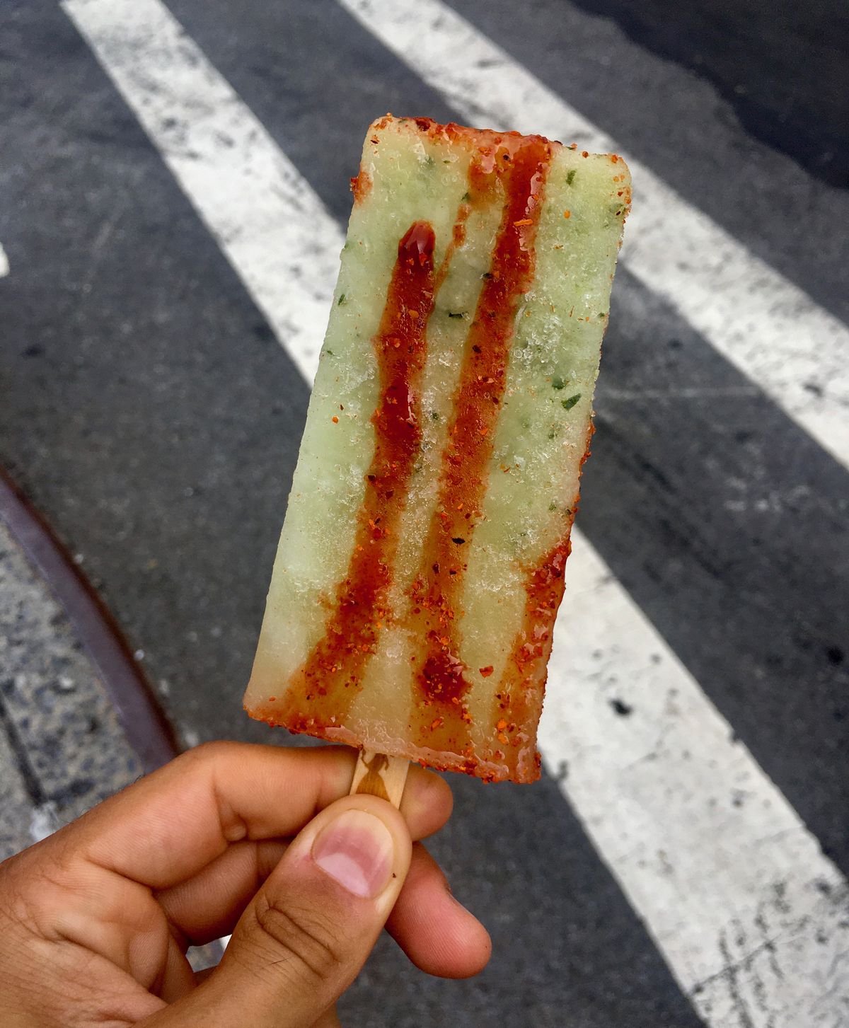 A cucumber lime frozen popsicle is covered in hot sauce and chile lime seasoning. In the background, a crosswalk is visible.
