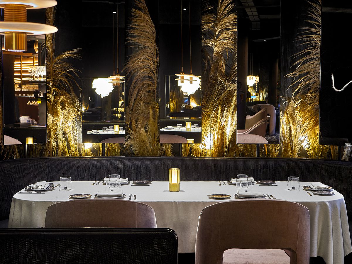 A dark restaurant exterior with gold lights illuminating dry wheat-like plants set between large mirrors, midcentury pendant lights, and a table set with white tablecloth and places for dinner. 