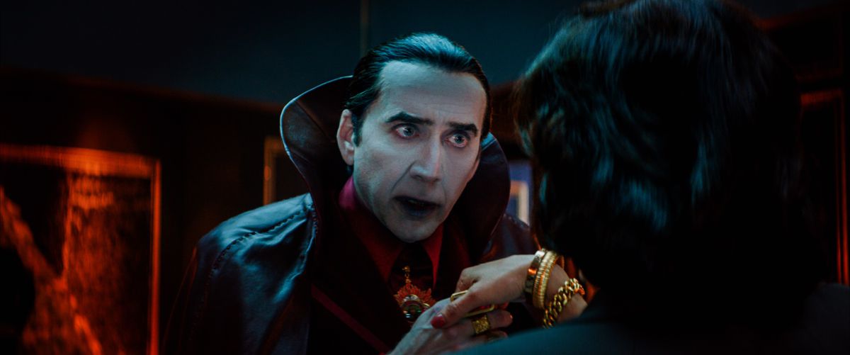 Nicolas Cage as a very traditional movie Count Dracula (pale-as-paper skin, slicked-back black hair with a widow’s peak, black cape with a huge high collar, hungry look in his eyes) takes the hand of a woman with her back to the camera in Renfield