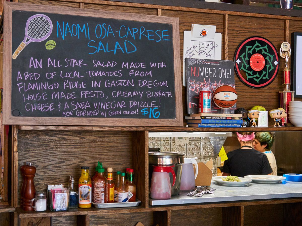 A chalkboard featuring a “Naomi Osa-Caprese Salad” special in front of the kitchen at The Sports Bra.