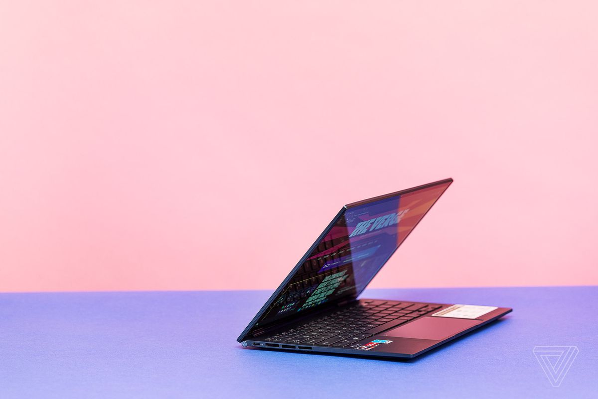 The Asus Zenbook S 13 OLED half open on a pink and blue background. The screen displays The Verge homepage.