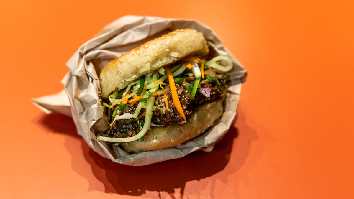 A sesame pancake sandwich in a takeout wrapper houses carrots, a mixture of darkened mushroom, and other accoutrements on an orange countertop