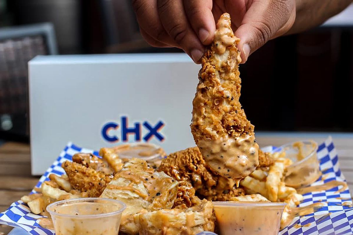 A hand dips a chicken finger in a rosy, peppery sauce. A basket with blue and white checked paper holds crispy chicken fingers and crinkle cut fries
