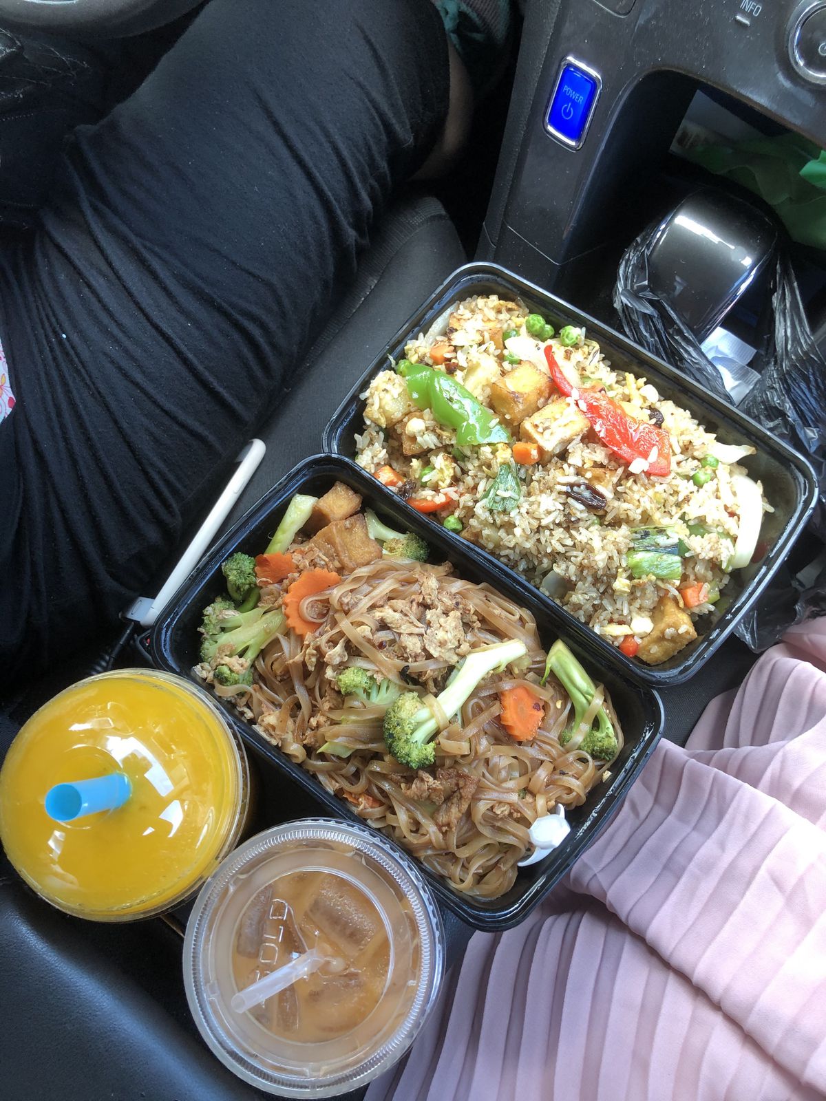 Top-down look at a two plastic takeout containers, one filled with pad Thai and one with fried rice, positioned on the booster seat of a car. Two beverages with straws sit in the cupholders.