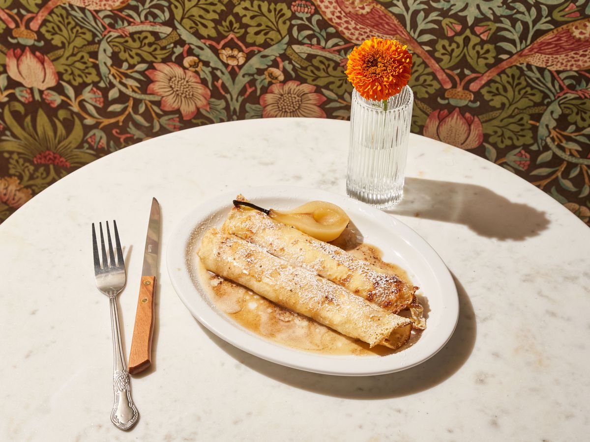 Two rolled crepes on a white plate sit side by side to a poached pear, atop a marble white table with silverware and a vase.