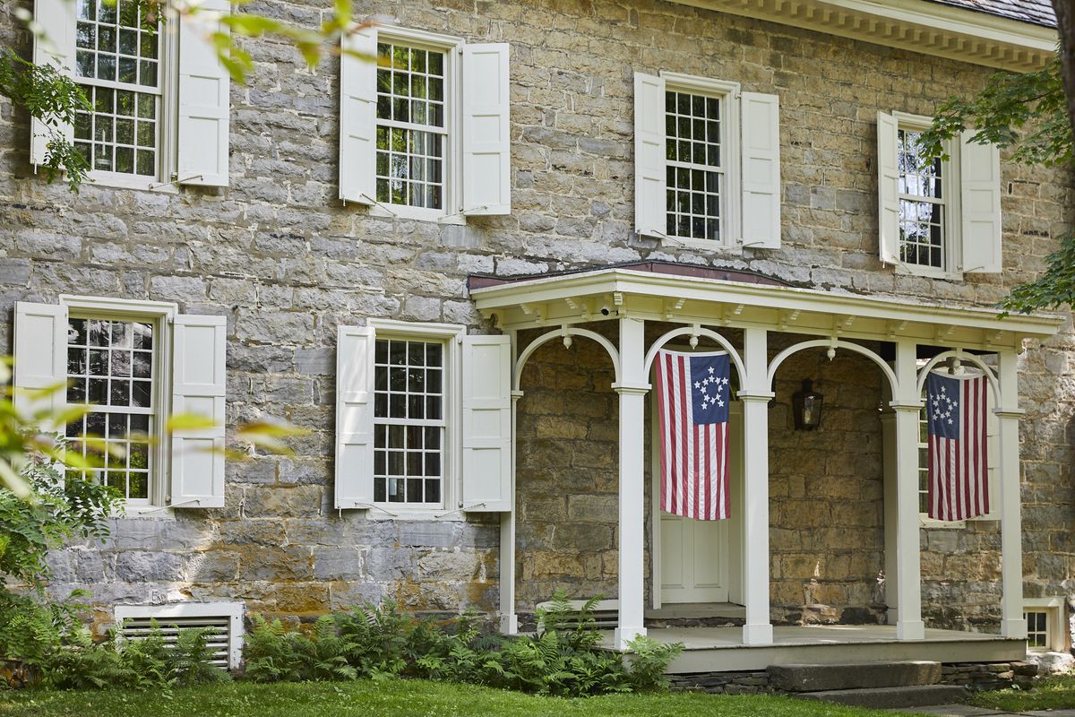 The exterior of a house. The facade is stone and there is a porch with hanging historic colonial United States flags.