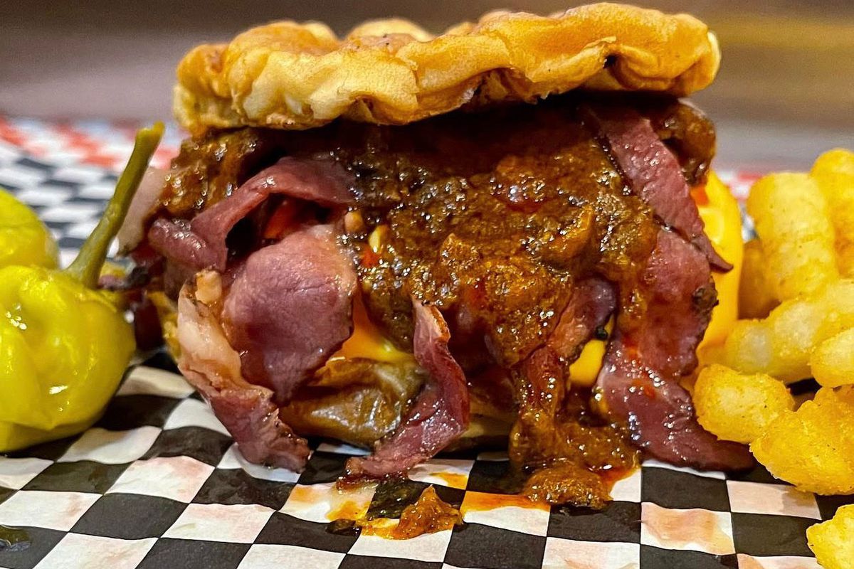 an extremely messy-looking chili cheeseburger topped with shaved pastrami and serves on B&amp;W checkered paper