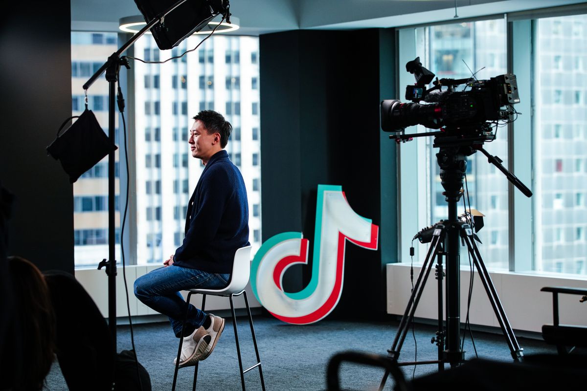 TikTok CEO Shou Zi Chew sits on a tall stool in a high-rise office backed by windows, surrounded by lights and cameras. 