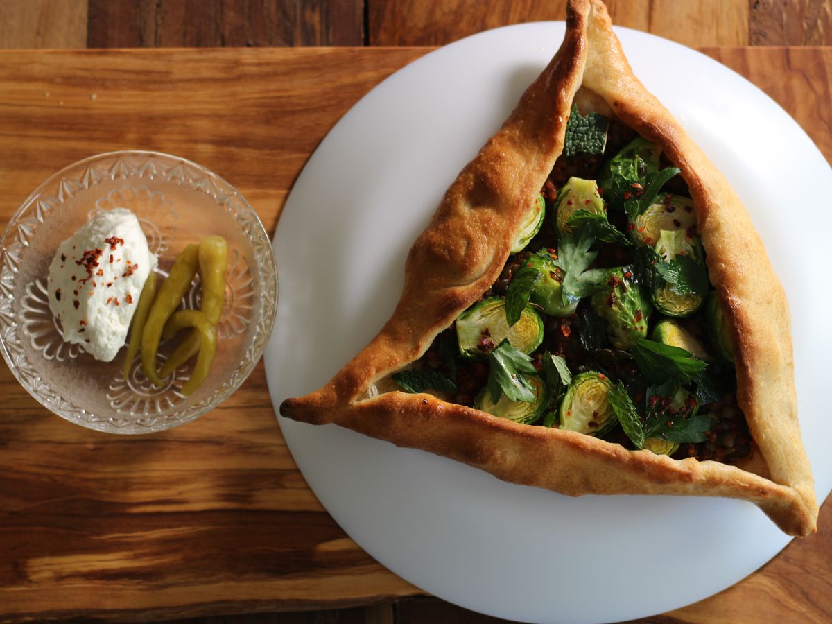 Overhead view of a triangular hand pie stuffed with Brussels sprouts. On the side, there’s a small clear bowl with a scoop of a thick white sauce and a pickled garnish.