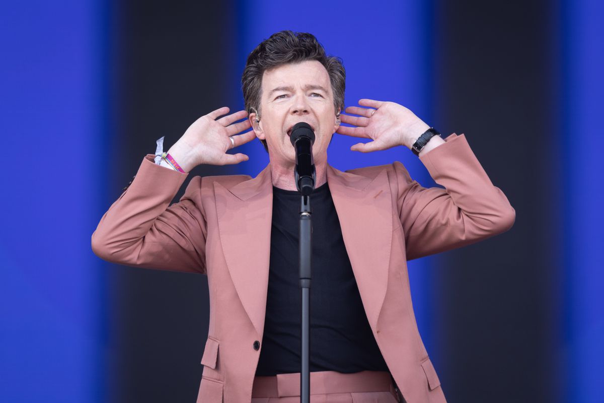 Rick Astley sings into a microphone with both hands raised near his ears. He wears a mauve suit jacket and pants and a black shirt. The performance took place at Worthy Farm, Pilton, in Glastonbury, England, on June 24, 2023.