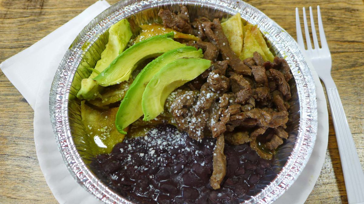 A round aluminum container with shreds of steak, beans, avocados and a few tortilla chips seen.