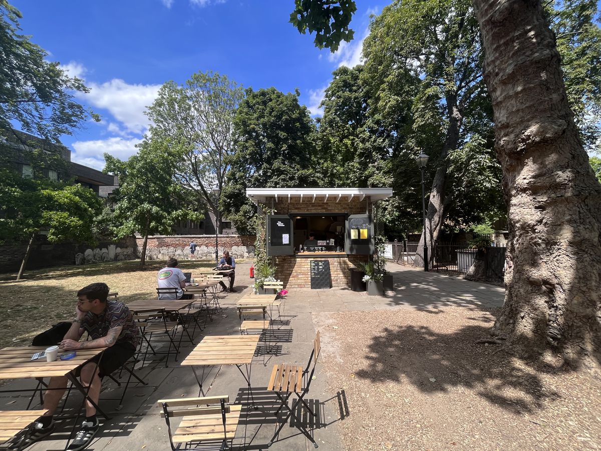 A garden with a small food kiosk at its rear, with tables and chairs scattered in front next to a tree.