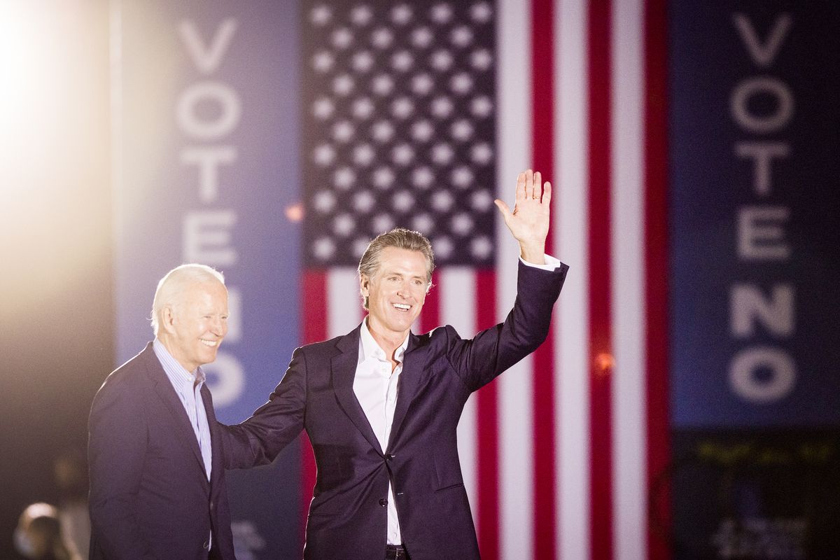 California governor Gavin Newsom waves from onstage with President Joe Biden in front of an American flag backdrop and signs that read, “Vote no.”