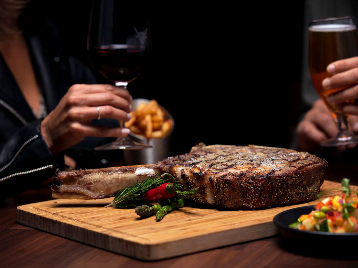 A steak on a wooden cutting board on a table in a dark dining room.