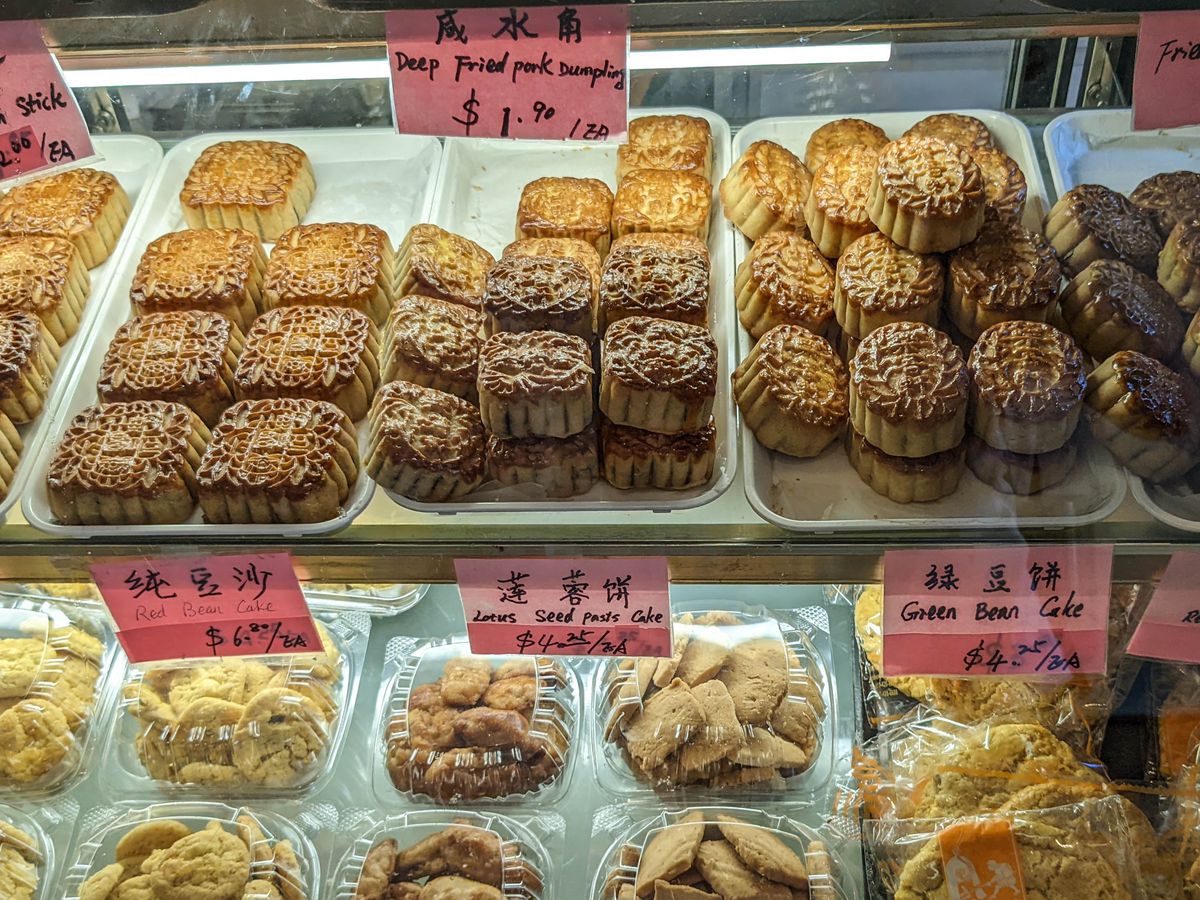Trays of golden-brown mooncakes with intricately shaped crusts are in a pastry case along with other Chinese pastries.
