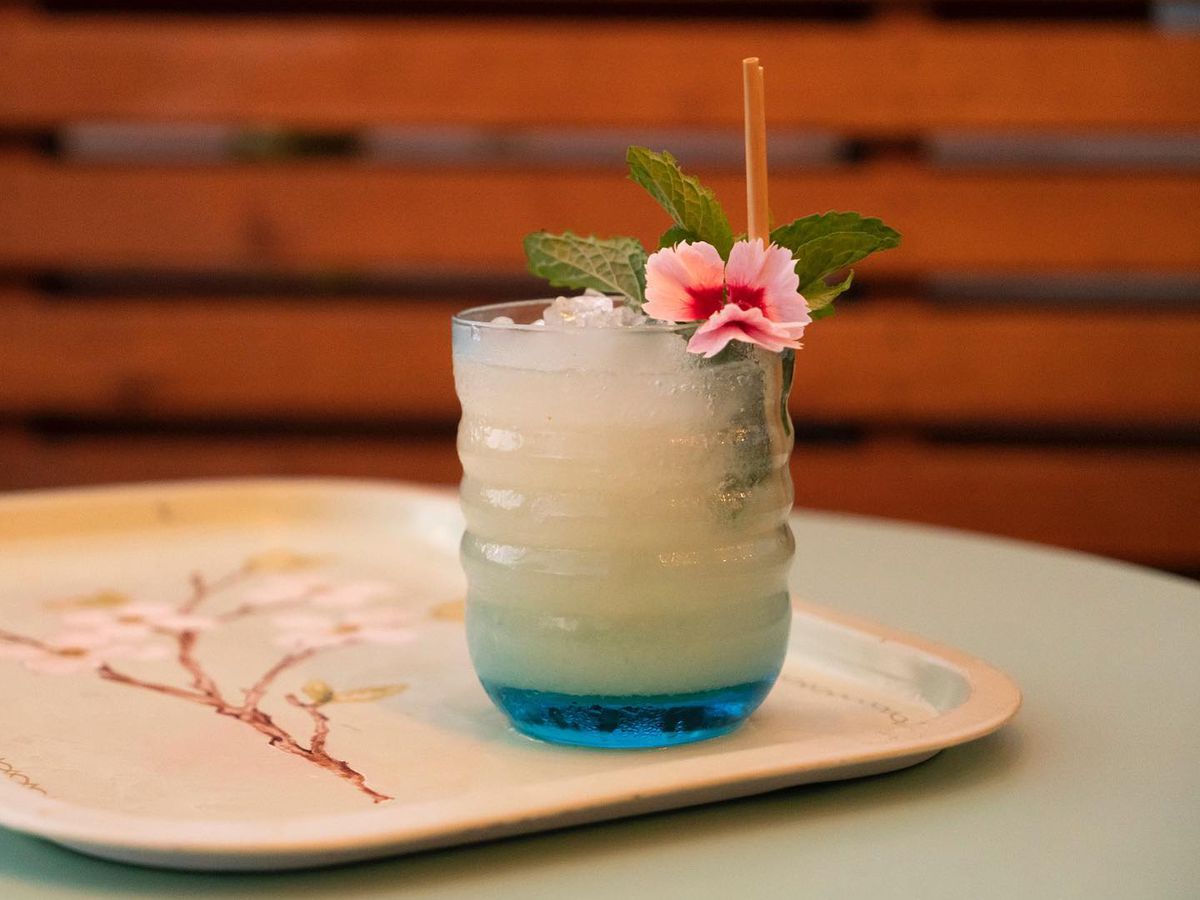 A ribbed glass with a cocktail over crushed ice, with a bright flower for garnish, sitting on a serving tray decorated with spindly branches, in front of wood slats blurred in the background