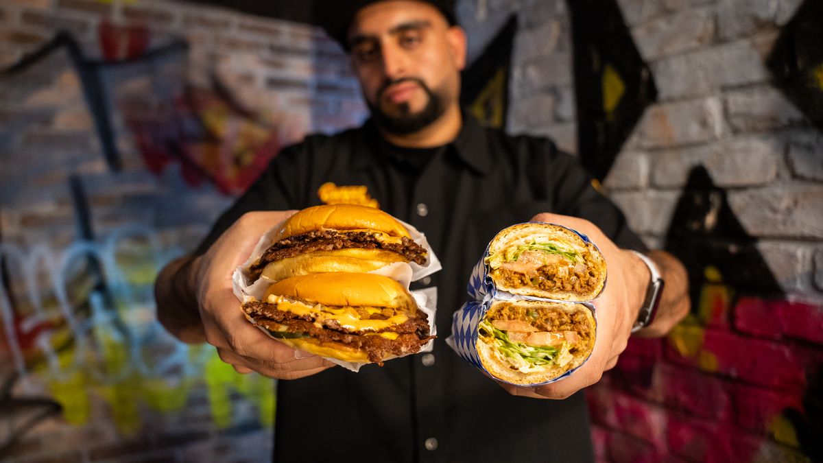 Burger Bodega Abbas Dhanani holding a smash burger and chopped cheese sandwich in front of a graffiti-ed wall.