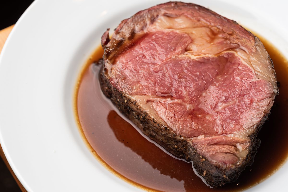 A thick cut of prime rib, pink on the inside, sits in jus on a white plate.