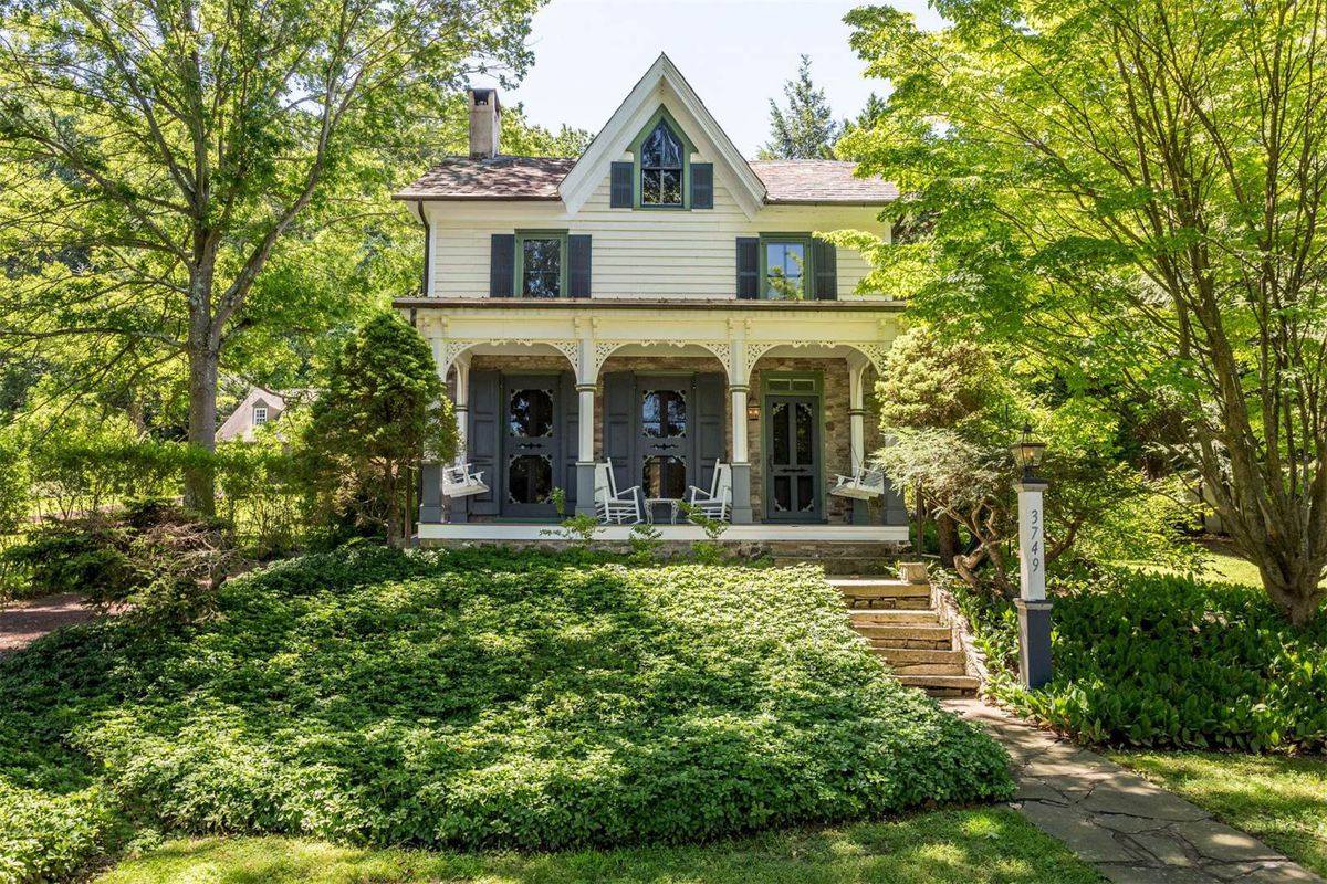 Two-story white home set back from the leafy street with covered front porch with ornate woodwork and green shutters and windows. 