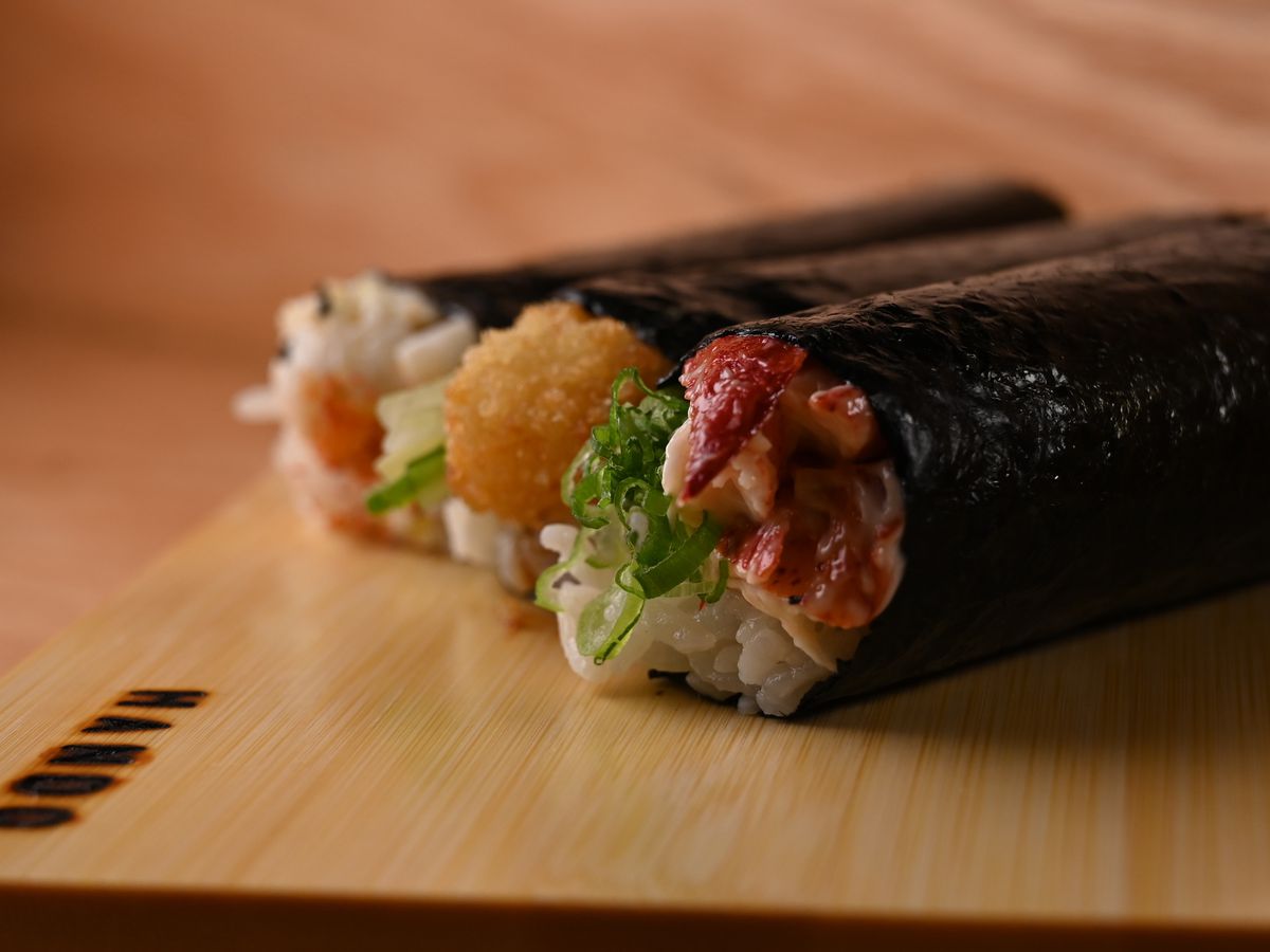 Three handrolls stuffed with an assortment of ingredients, sitting atop a wooden plank.