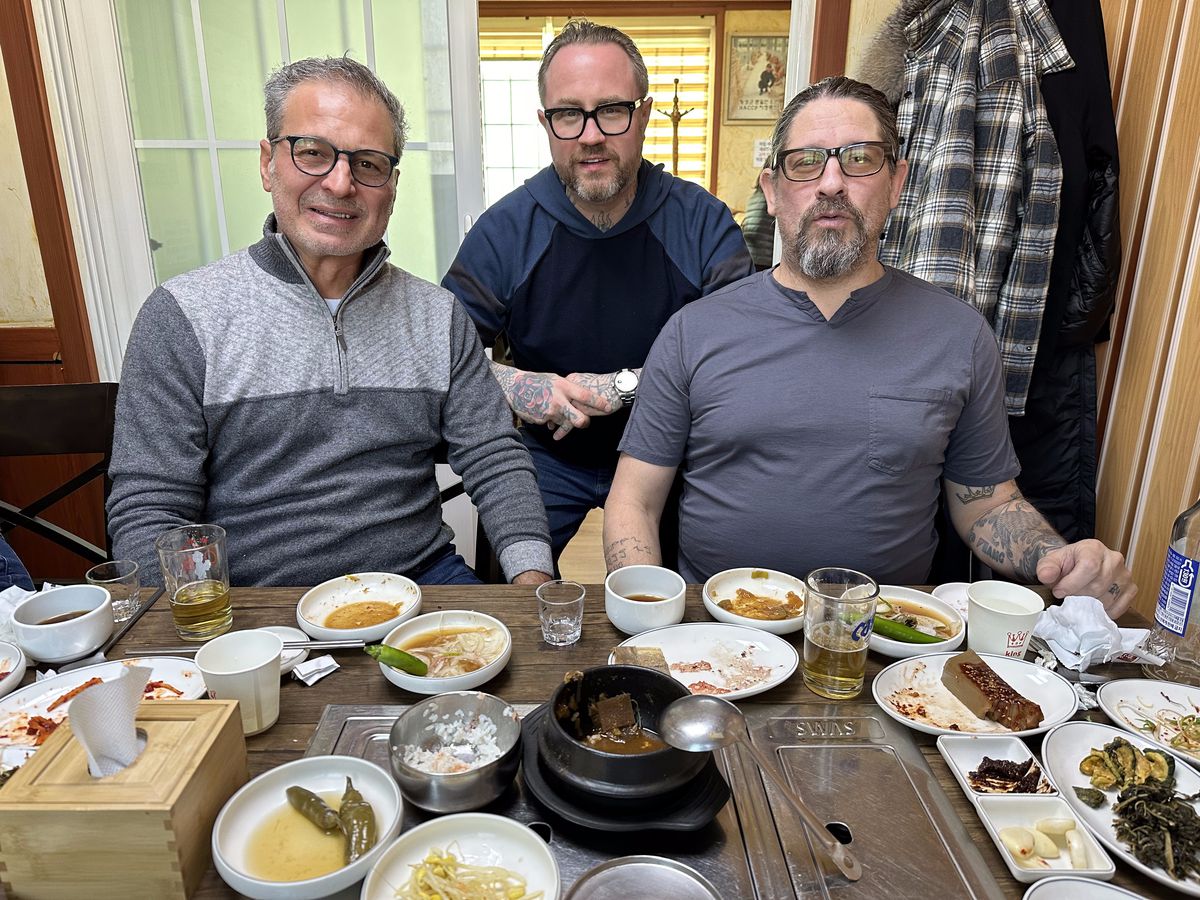 Three men pose for a photo over a restaurant table covered in plates and bowls after a meal.