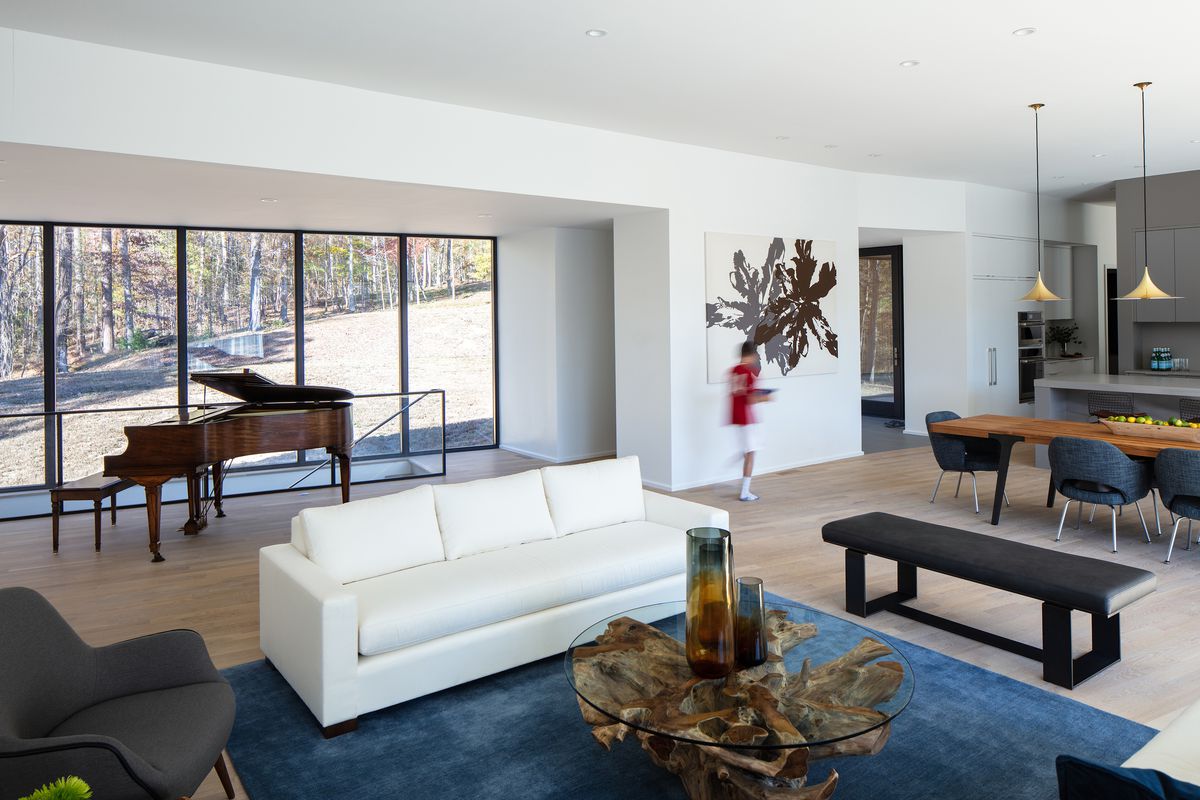 A living room. There is a white couch and a blue area rug. The floor is hardwood. There is a piano, table, multiple chairs, and a bench. A large work of art hangs on the wall.