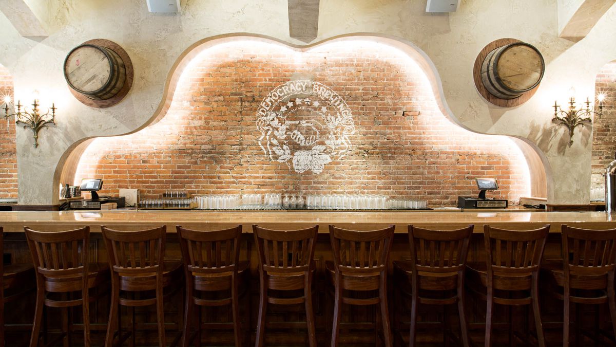 A wooden bar with an illuminated brick wall in the rear and two half barrels hanging overhead. A logo painted on the wall reads “democracy brewing”