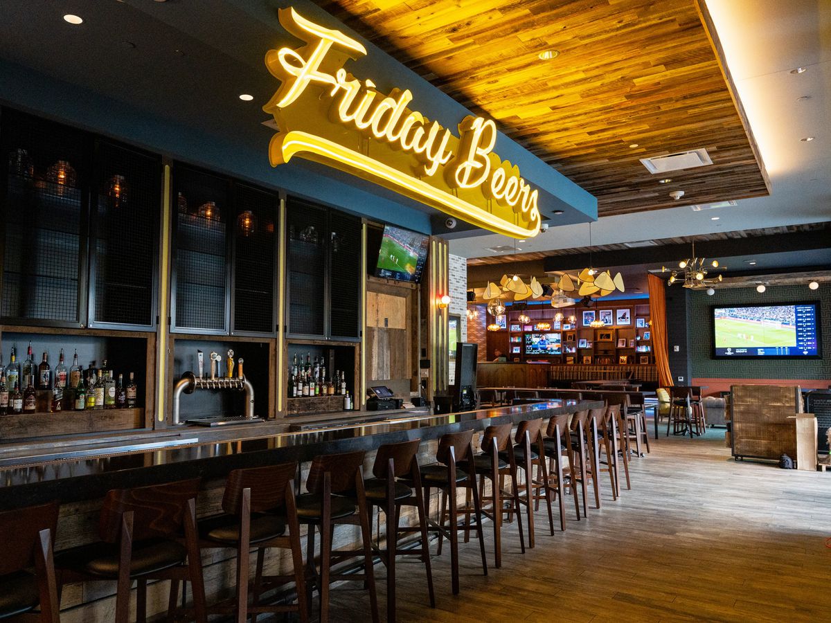 A long wooden bar lined with empty bar chairs with a sign above it that says “Friday Beers” in yellow cursive. In the back are tables and chairs with a big screen TV