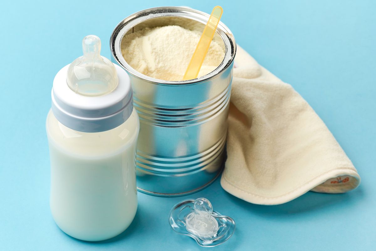 Open can of baby formula with a scoop inside of it next to a bottle filled with white liquid and a pacifier.