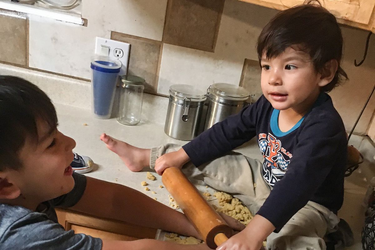 A 2-year-old sits on the counter holding a rolling pin while his 5-year-old brother helps him roll out buñuelos at home.