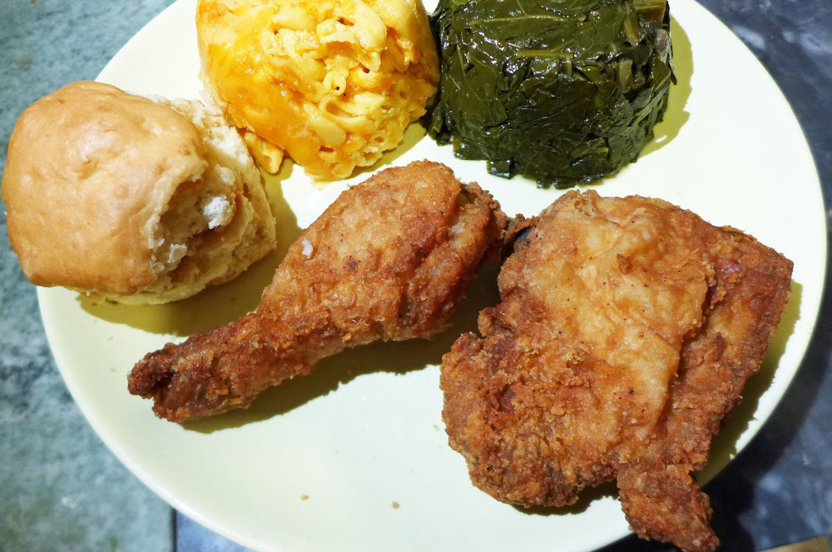 Two pieces of fried chicken on a white plate with collards, mac and cheese, and a biscuit