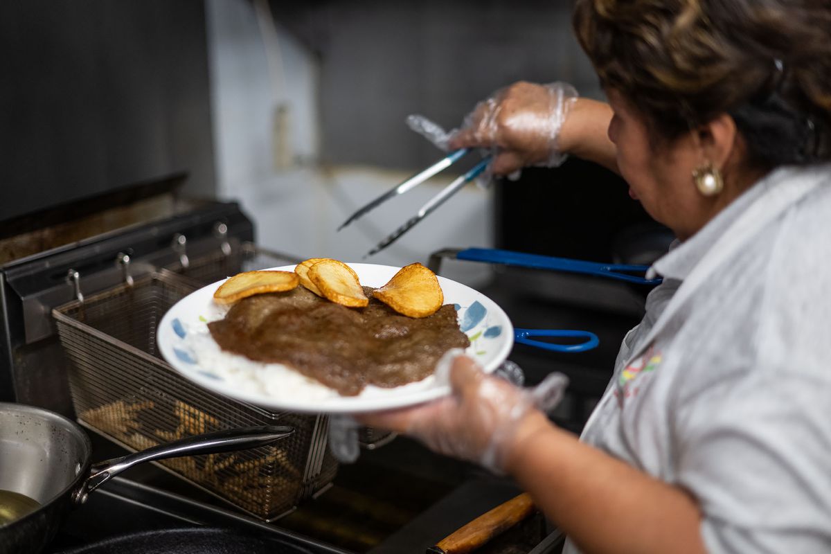 A woman plating a griddled meat with fried potatoes inside a restaurant kitchen.