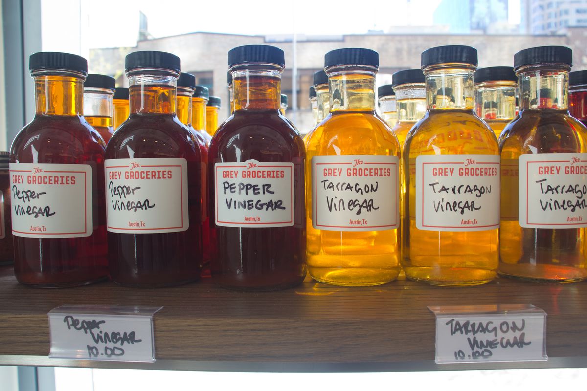 Bottles of red liquid with labels reading “Grey Groceries: Pepper Vinegar” next to bottles of yellow liquid with labels reading “Grey Groceries: Tarragon Vinegar” on a shelf. 
