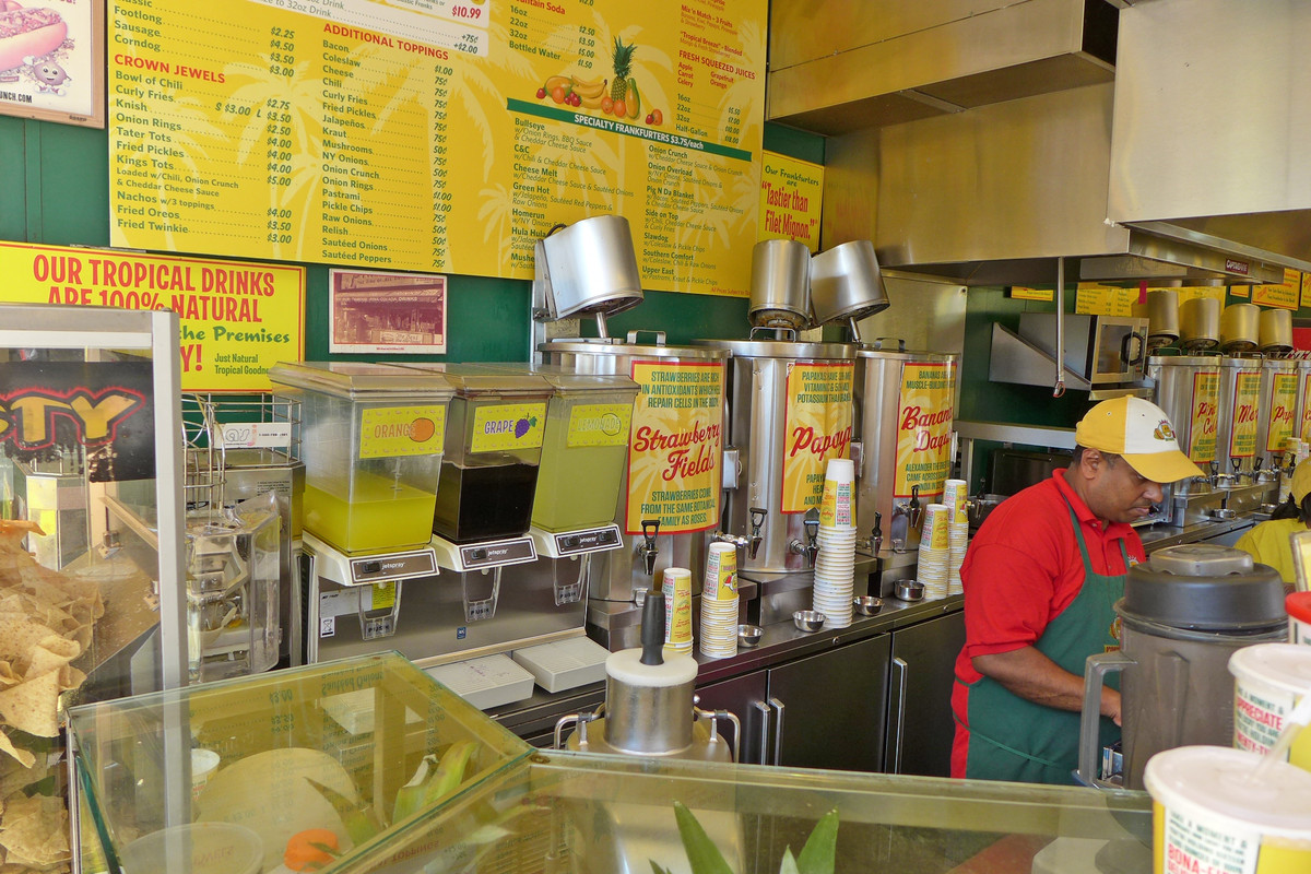 Papaya King’s Upper East Side location is covered in yellow menu signs as an employee works at right of frame.