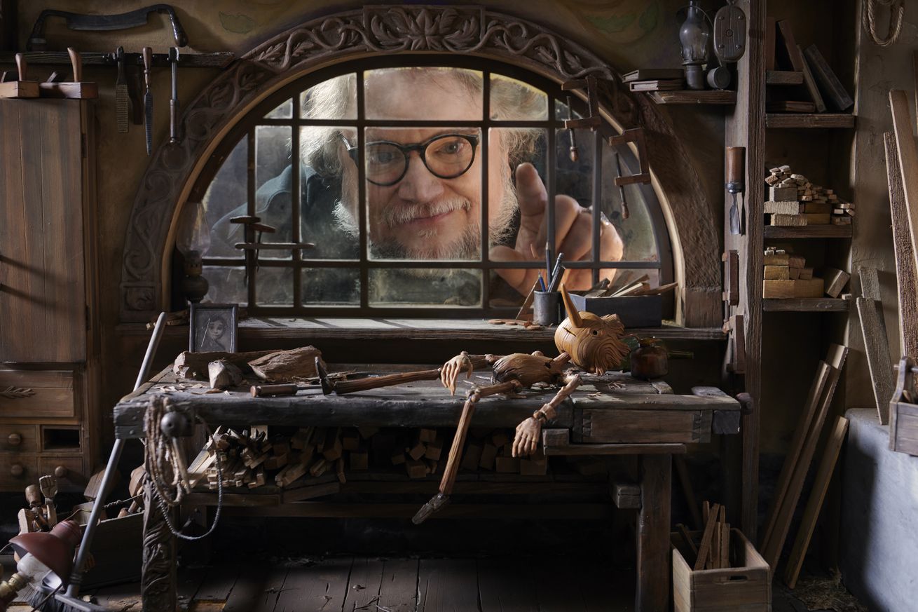 A miniature scene depicting Pinocchio lying in Geppetto’s cluttered workshop as Guillermo del Toro looks on through a window in the background.