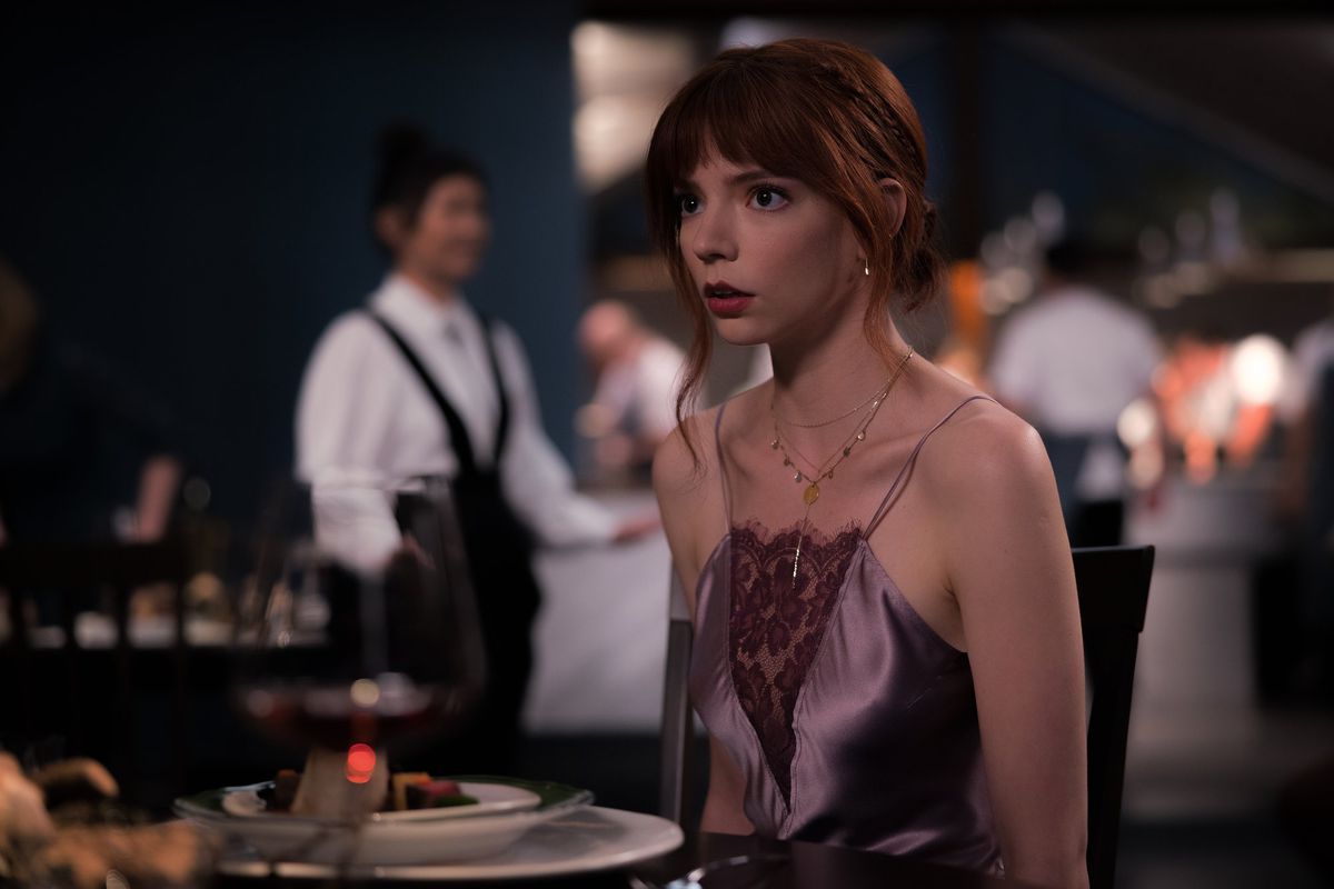 A red-haired woman in a purple dress (Anya Taylor-Joy) sits at a table with an open-mouthed expression of worry on her face.