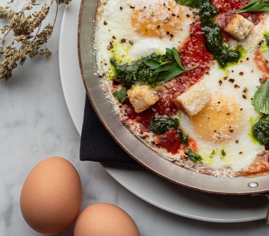 A partial overhead picture of sunny side up eggs in a circular dish filled with red tomato sauce, green basil leaves and croutons. A couple of uncracked brown eggs and sprig of dried herbs sit beside the dish on a white marble counter.