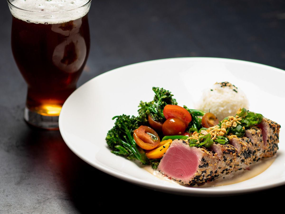 A sesame crusted seared ahi tuna, served with vegetables and steamed white rice. All sits on a white plate, over a dark grey table. A small glass of cuvee beer is on the side.