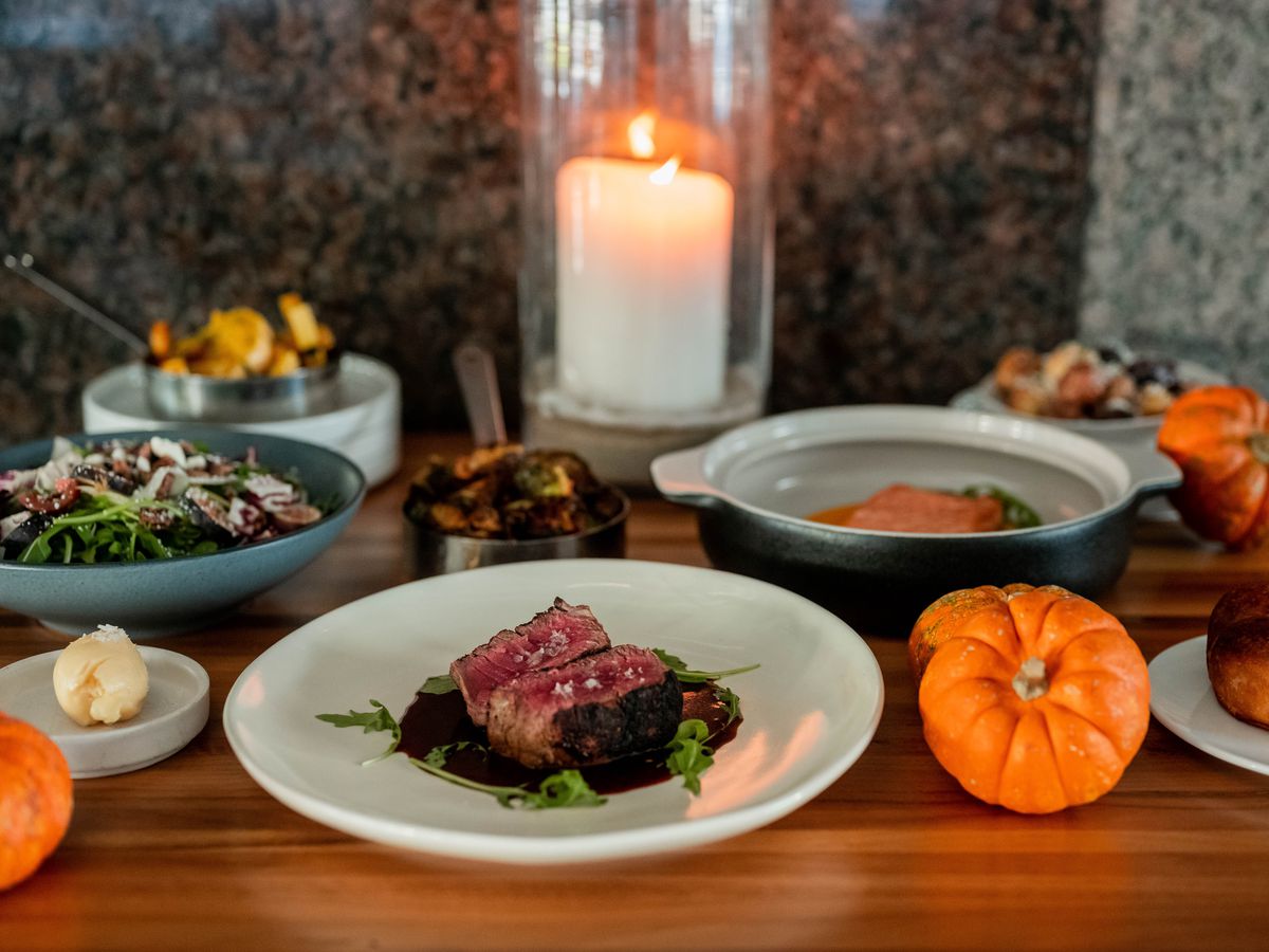 A table set with fall decor holds a burning white candle, a plate of rare steak, a green salad, and yams.