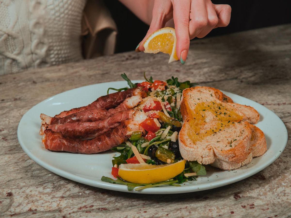 An image depicting a food dish consisting of prosciutto-wrapped shrimp, greens, and bread with a hand squeezing a lemon over the food.