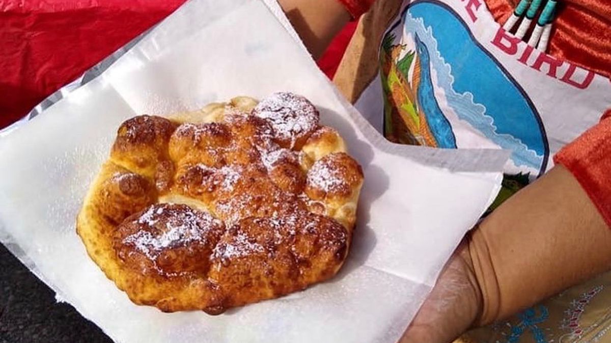 Hands hold out a square piece of parchment with a brown and bubbly piece of frybread on top. 