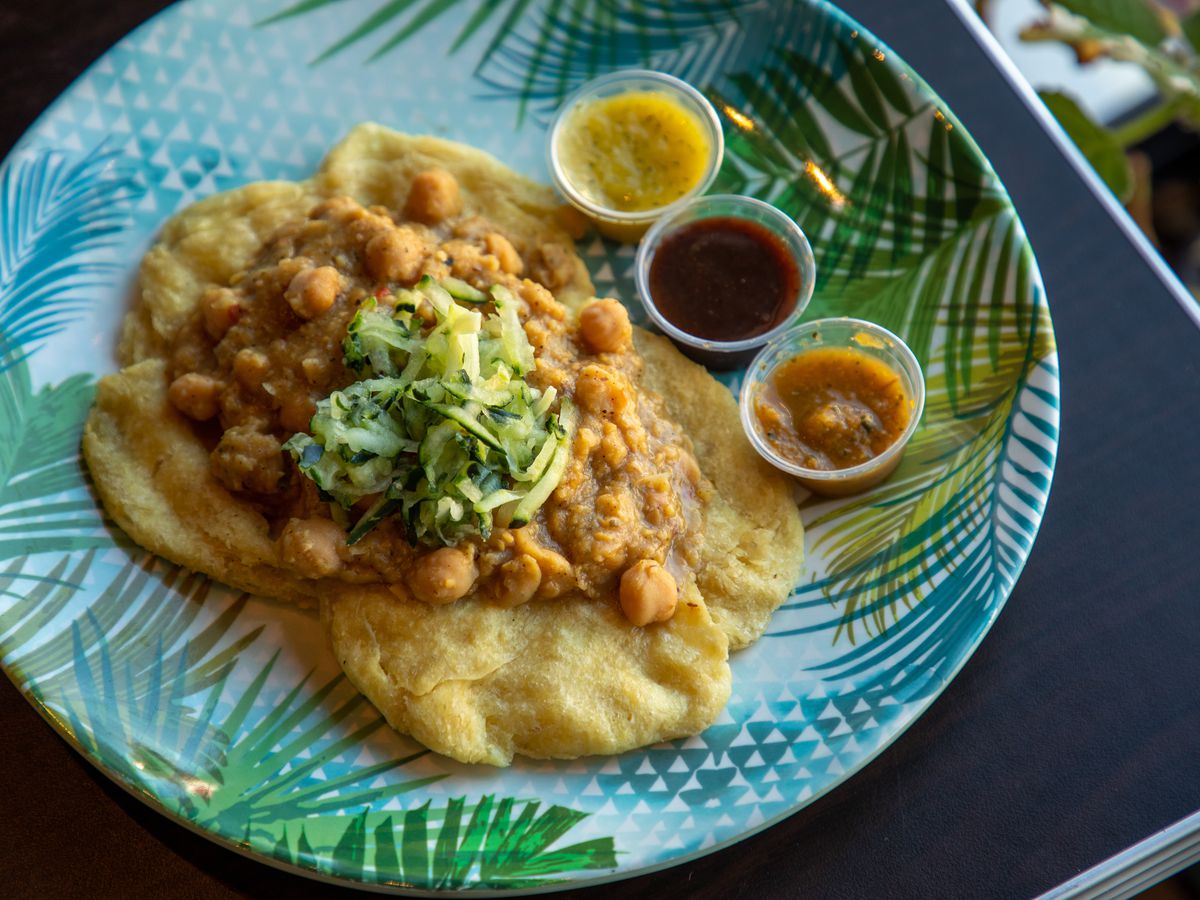 A colorful plate of two pieces of fried flat bread topped with a chickpea filling and cucumber salad next to three sauces.