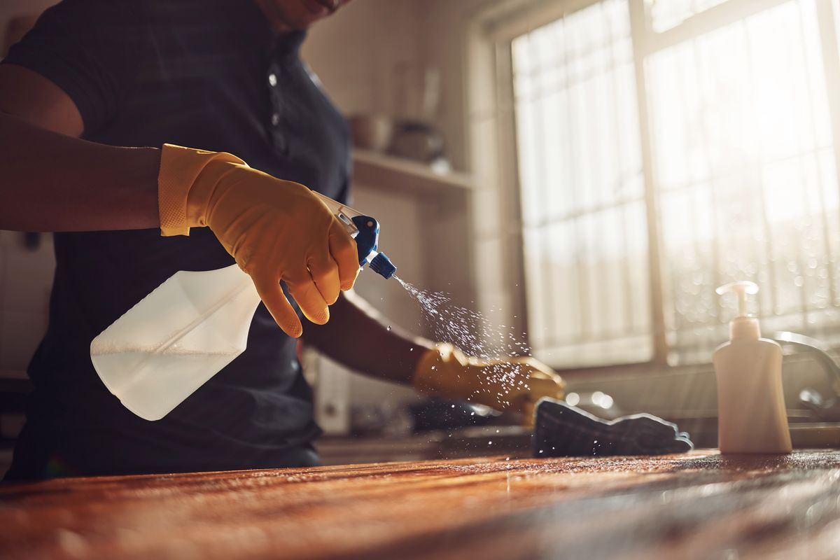 A woman cleans a butcher block countertop with a spray bottle and gloves. 