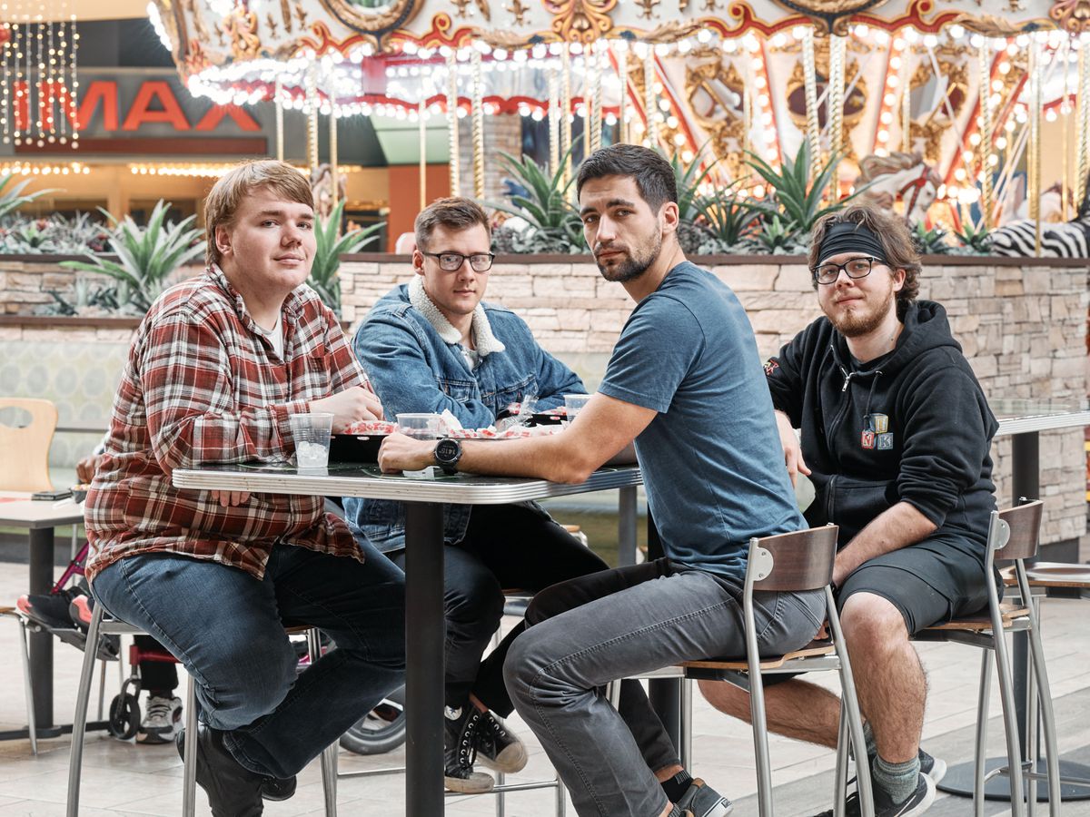 Four young men sitting around a food court table with a lit-up carousel in the background.