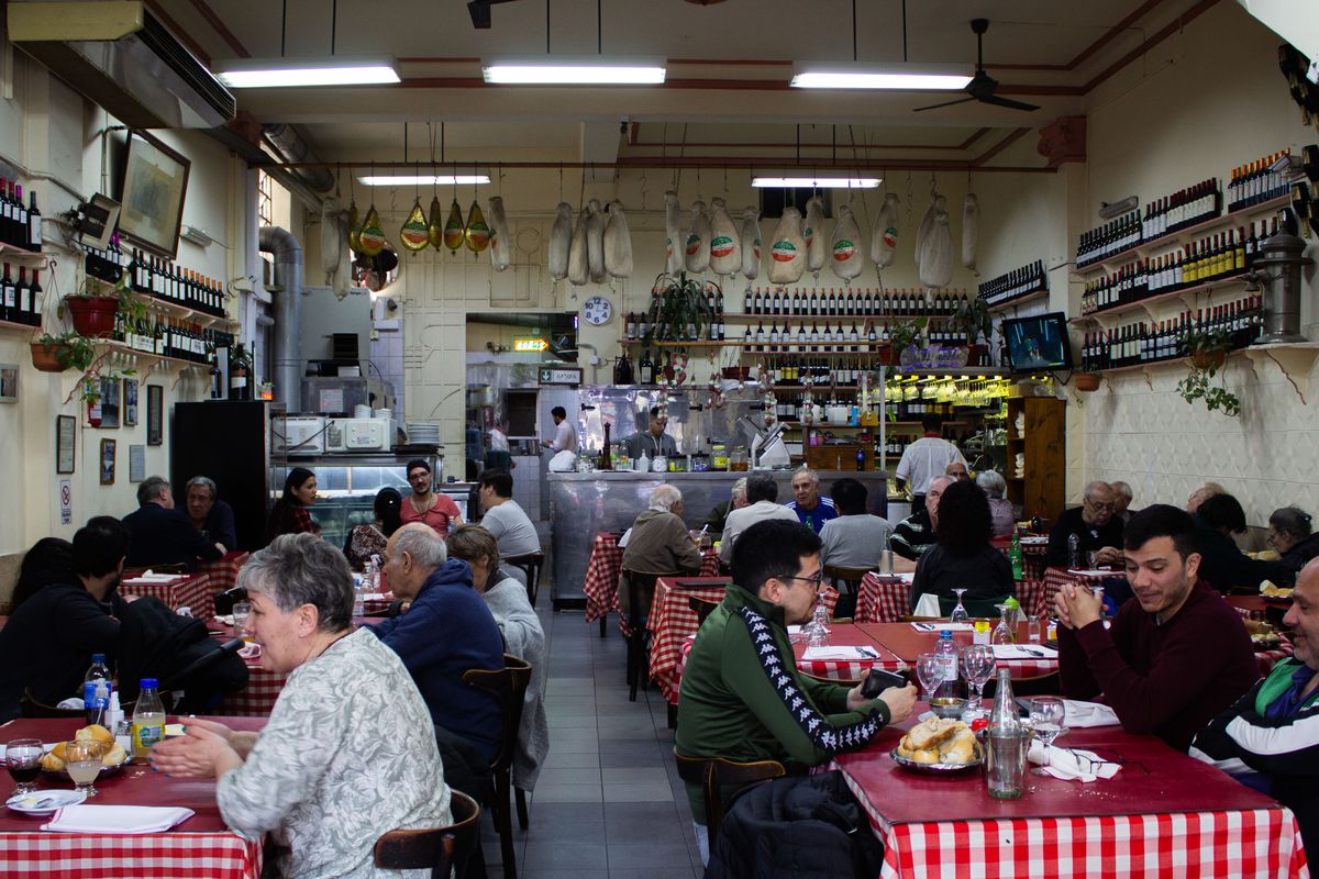 A packed restaurant interior with diners at red checkered tablecloths, hunks of meat hanging from the ceiling, cooks working behind a counter in the back, and rows of wine bottles on the walls.