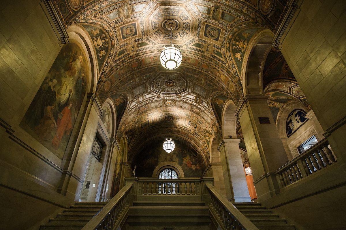 The interior of the Detroit Public Library. There is a large staircase leading to an upper level. The ceiling is elaborately decorated with an intricate design. There are large paintings and archways on both sides of the staircase.