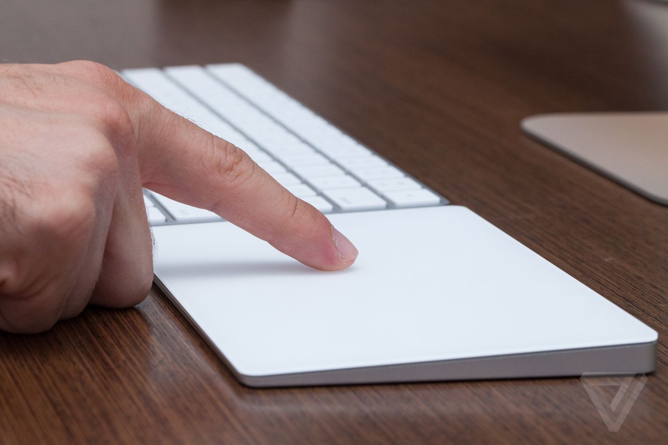 A finger touching Apple’s Magic Trackpad, which sits next to a keyboard on a desk.